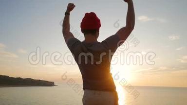 徒步旅行者站在靠近大海的悬崖边，举起双手迎接令人惊叹的日落。 快乐的人到了