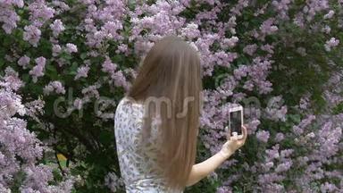 一个年轻漂亮的女孩正在欣赏一丛美丽的丁香花。 在智能手机上自拍。 夏日，好心情..