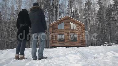 幸福的年轻夫妇站在木屋旁。 库存。 冬天，一对夫妇站在森林里的房子附近
