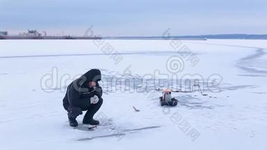 穿着深色衣服的老渔夫在河港的背景下在冰冻的河面上用冬季的鱼竿钓鱼