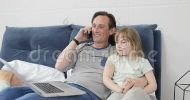 父亲拥抱小女儿，一边打电话，一边快乐地微笑着，一家人坐在床上