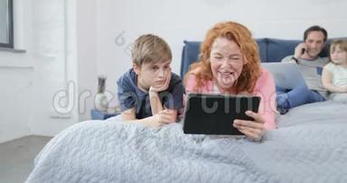 一家人一起在卧室里度过一个早晨母亲和儿子一起看视频，嘲笑父亲在打电话