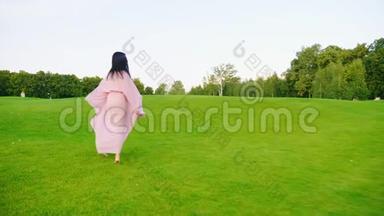 穿着淡粉色裙子的孕妇。 穿过绿色的草地