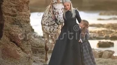妈妈和女儿穿着优雅的衣服，在阳光明媚的阳光下，带着美丽的斑点马走在岩石海岸上