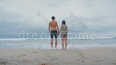 年轻的一家人牵着手站在海滩的海洋前。 旅<strong>途中</strong>享受海浪的年轻夫妇