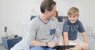 父亲和儿子在卧室里拿着<strong>数码</strong>平板电脑坐在床上，会说话的<strong>家</strong>庭在卧室里