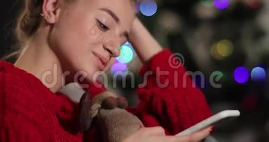 新年庆典。 穿着红色毛衣的迷人年轻<strong>女子坐在</strong>圣诞树前的<strong>沙发上</strong>，和她聊天