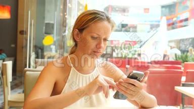 一个金发女人正坐在一个炎热的夏天的咖啡馆里，微笑着在手机上输入短信。 4k.慢
