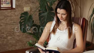 年轻漂亮的女孩坐在椅子上看书。 迷人的女人一边看书一边微笑。 积极的情绪