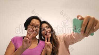 退休母亲和年轻女儿展示和平标志和制作有趣的自拍照片与手机。 4K.