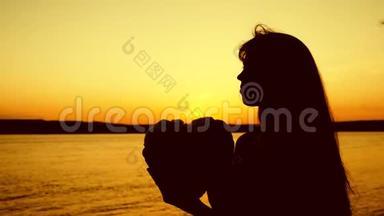 在美丽的夕阳下，女孩的剪影紧紧地抓住大海。 美丽的女孩在夕阳下抱着心。