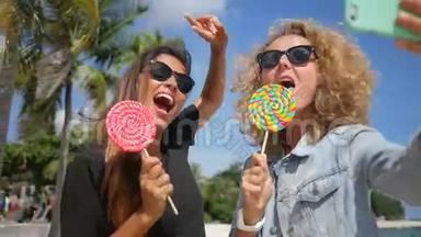 两个年轻的少女`在热带海滩用手机和舔棒棒糖做自拍。 高清慢速运动。 苏