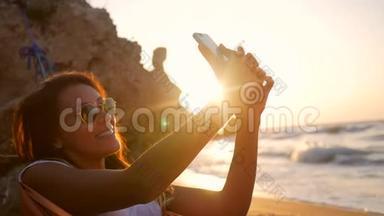 年轻的混合种族女孩拍摄自拍照片，同时放松在海滩上的吊床在惊人的日落。 高清慢速运动。
