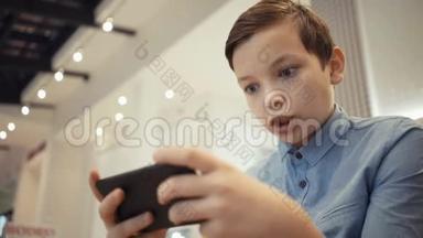可爱的男孩坐在鼹鼠大厅的长凳上，在智能手机上的游戏中情绪化地玩