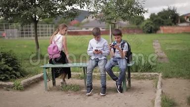 两个男孩和女孩在放学后使用他们的电话。 可爱的男孩坐在长凳上玩网络游戏