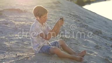 戴着耳机的男孩在智能手机上玩游戏。 10岁的孩子在日落时分坐在沙滩上享受智能手机。