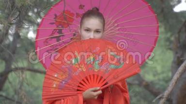 一个穿着红色和服的艺妓，她的肩膀上有一把伞，用一把漂亮的扇子遮住她的脸，看着她。