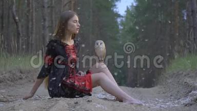 一个留着长发的女孩坐在森林中央的一条小路上，穿着一件黑色的衣服，看着一只浅棕色的猫头鹰