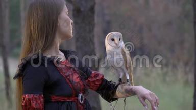 一个穿着黑衣服、有纹身的女孩正在检查一只浅棕色的猫头鹰，她坐在手臂上，在森林里射击，有点慢