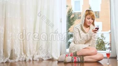 穿着毛衣的漂亮女人坐在靠近窗户的地板上，在电话上<strong>发信息</strong>