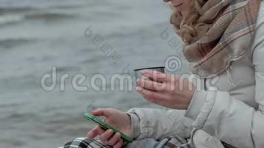 年轻女子穿着暖和的衣服坐在海边，坐在木盘管上，用电话从<strong>热水</strong>瓶里<strong>喝</strong>热茶
