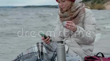 年轻女子穿着暖和的衣服坐在海边，坐在木盘管上，用电话从<strong>热水</strong>瓶里<strong>喝</strong>热茶