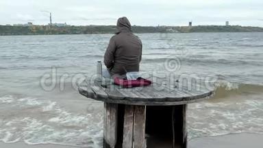 穿暖和衣服的年轻人坐在海边，坐在木盘管上，用<strong>热水</strong>瓶<strong>喝</strong>热茶