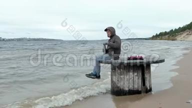 穿暖和衣服的年轻人坐在海边，坐在木盘管上，用<strong>热水</strong>瓶<strong>喝</strong>热茶