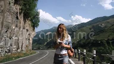 一个穿裙子的年轻金发旅游女孩正走在山里的路上，看着电话