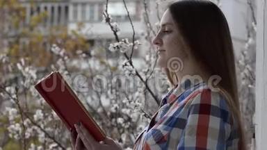 穿着格子衬衫的女孩站在<strong>窗边</strong>的阳台上，读着一本关于盛开的杏树<strong>背景</strong>的书