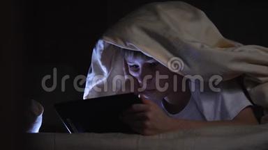 一个孩子晚上在毯子下面使用平板电脑。 这个男孩玩电脑游戏