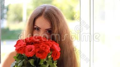 一个年轻的女人，在一扇开着的窗户拿着一束红玫瑰。 一个美丽的女孩喜欢花香