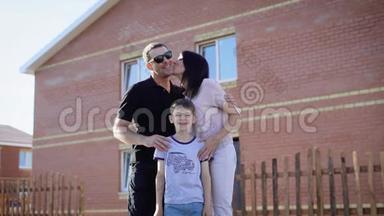 幸福的家庭站在他们的房子附近，在阳光明媚的夏天互相拥抱。 父亲、母亲和儿子感到高兴