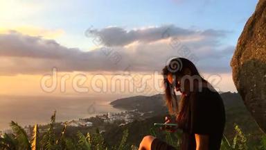 美丽的女人坐在悬崖上，在智能手机上发短信。 泰国普吉岛。 高清慢动作惊人的视野。