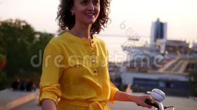 一个身穿黄色长裙的微笑着的黑发女人站在她的城市自行<strong>车</strong>旁，<strong>车把手</strong>里拿着鲜花