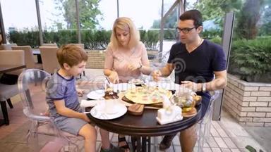 父亲和母亲和儿子坐在餐桌旁，在餐馆里给儿子一片披萨