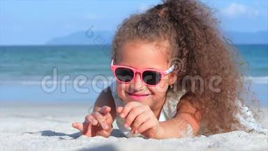 一个戴着粉红色眼镜的漂亮小女孩的特写肖像，可爱的微笑着看着相机，躺在沙滩上。