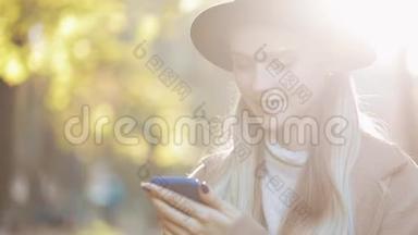 年轻女子穿着外套，用智能<strong>手机</strong>站在秋天公园。 户外<strong>科技</strong>。 阳光