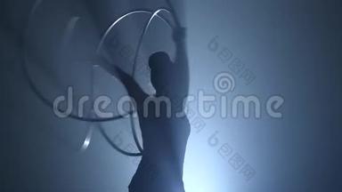 空中体操运动员用双手拧动金属圈。 慢动作。 烟雾背景。 剪影