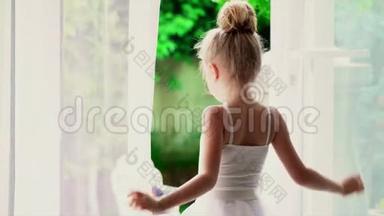 美丽的小芭蕾舞女看着窗外的大自然。 穿着白色短裙的小女孩在窗边