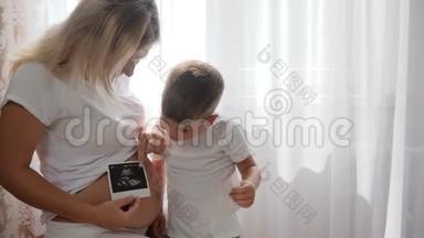 年轻的怀孕妈妈，带着甜美的孩子，在明亮的自然光线下在家里看摄影声像