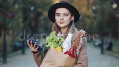 年轻漂亮的女人穿着时髦的外套，站在秋天的公园里，拿着一包产品，用智能手机