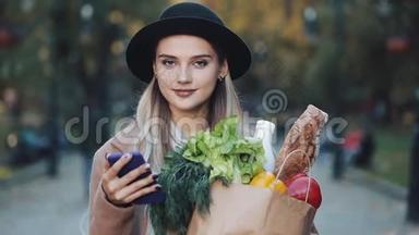 年轻漂亮的女人穿着时髦的外套，站在秋天的公园里，拿着一包产品，用智能手机
