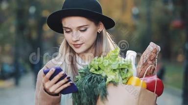 年轻漂亮的女人穿着时髦的外套，站在秋天的公园里，拿着<strong>一包</strong>产品，用智能手机