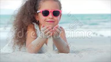 粉色眼镜中美丽的小女孩的特写镜头，可爱的微笑看着镜头，躺在沙滩上
