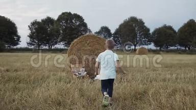 一个小男孩跑到一个女孩面前，她坐在稻草堆下的田野里。 快乐的女孩穿着白色的衣服，微笑的男孩奔跑
