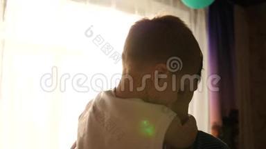 年轻的父亲抱着他的孩子靠近窗户。 阳光透过窗户。 孩子的欢笑和喜悦..