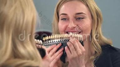一个金发女孩看着镜子前面的结果牙齿美白在一个特殊的颜色尺度。 女孩很高兴