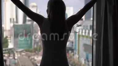 女人揭开窗帘的剪影，看着窗外，伸展着。 城外摩天大楼景观