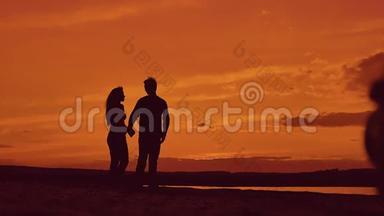 日落时分，一对夫妇在海滩上牵手散步的剪影。 在夕阳的灯光下，在海滨快乐的男人和女孩